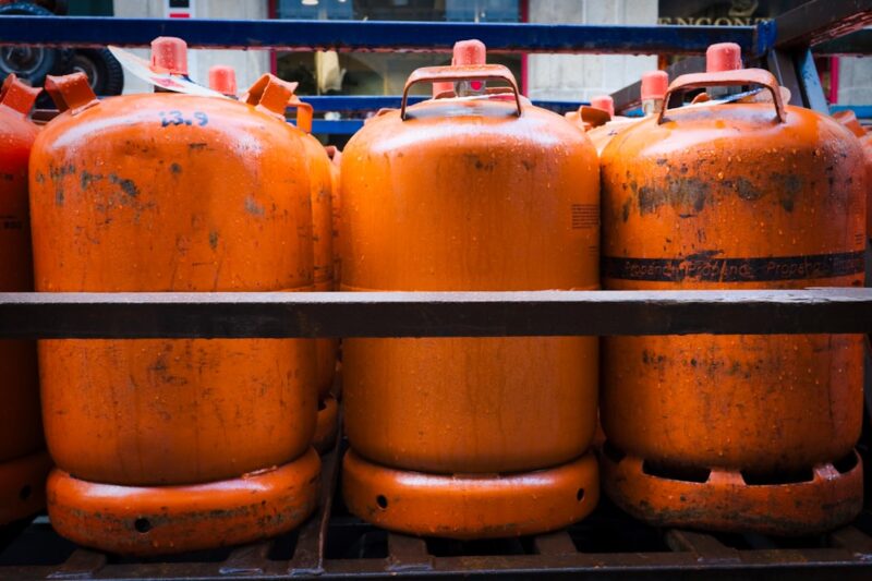 Volle gasfles inleveren op de milieustraat of bij leverancier Orange Propane Tanks Stacked on Metal Rack in City Setting - A close-up shot of three large orange propane tanks stacked on a metal rack in an urban setting. The tanks are in a row, and the rack appears to be part of a truck or loading dock. The tanks are worn and faded with evidence of rust and water droplets.