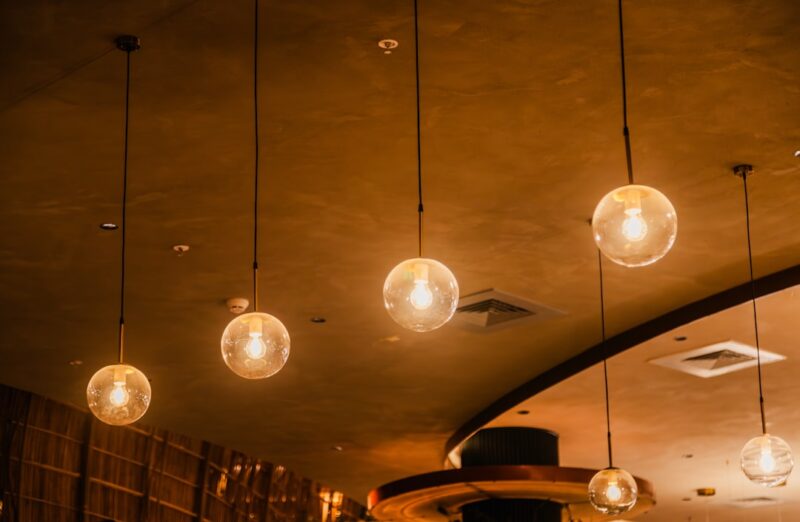 Verlichting ophangen zelf doen zonder risico met dit stappenplan The image displays several pendant lights hanging from the ceiling, each featuring a single light bulb encased in a clear, spherical glass globe. The lights emit a warm glow, creating an inviting and cozy ambiance. The ceiling is finished with a textured surface, adding to the modern and elegant interior design. The arrangement of the lights and their soft illumination enhance the overall aesthetic of the space.